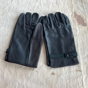 May include: A pair of black leather gloves. Each glove has a wrist strap with a buckle. The gloves are lying on a light-coloured, textured surface.