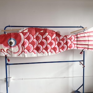 May include: A large, horizontal carp-shaped windsock, known as a koinobori, featuring a red and white scale pattern with a prominent eye. The fabric fish is suspended from a blue metal garment rack using wooden hangers and metal clips.