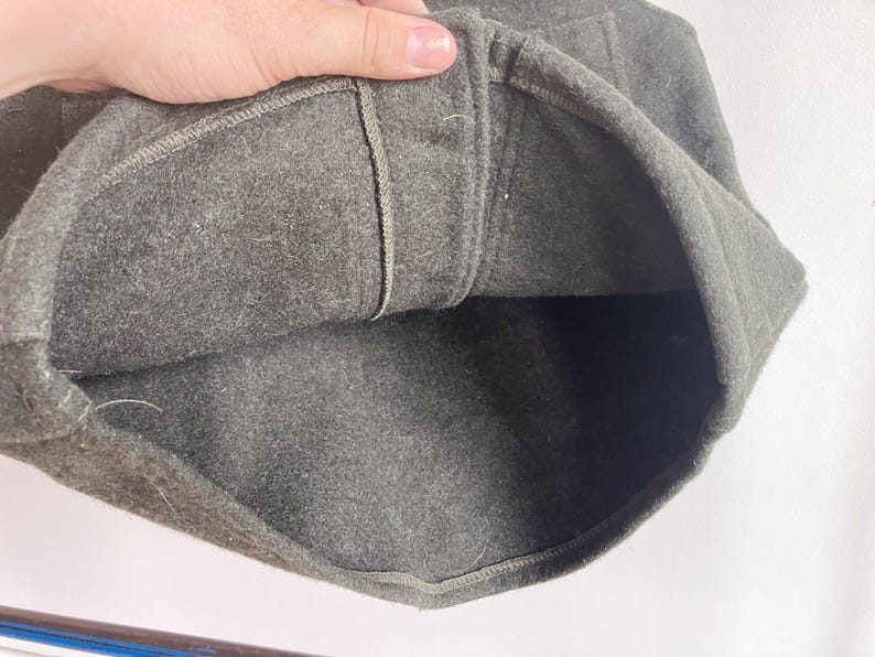 May include: A dark olive green felted wool cap featuring a structured crown and a thick, folded brim. The interior shows a matching dark olive green felt lining with visible stitched seams along the inner band and a reinforced top panel.