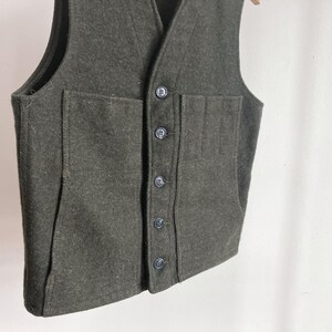 May include: A dark olive green vest featuring a V-neckline and a front button closure with five matching buttons. The garment includes two large patch pockets on the front, with the left pocket divided into three vertical sections for storage.