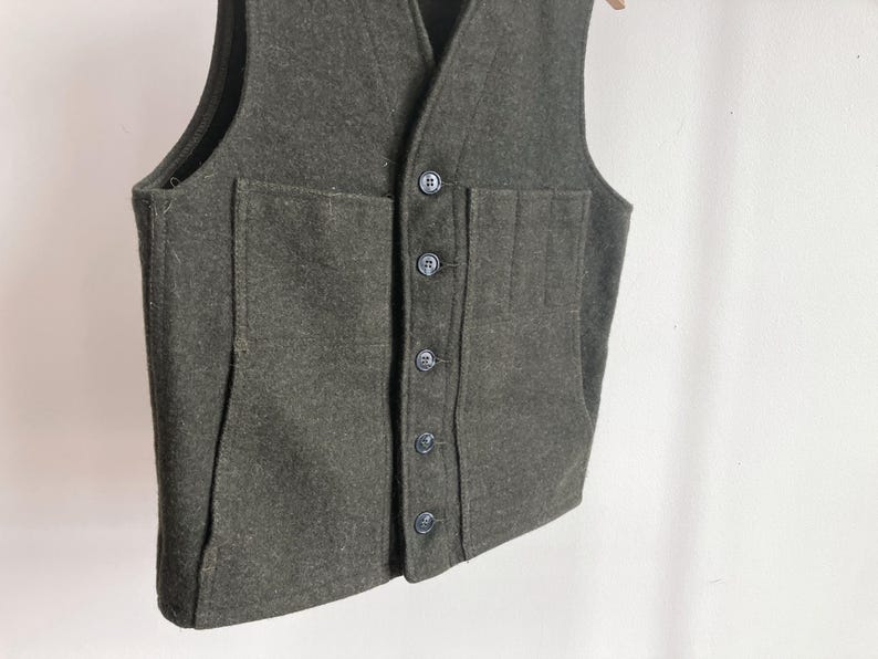 May include: A dark olive green vest featuring a V-neckline and a front button closure with five matching buttons. The garment includes two large patch pockets on the front, with the left pocket divided into three vertical sections for storage.