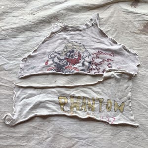 May include: Cream-coloured, distressed crop top featuring a cartoon character graphic with red accents. The bottom section displays the word "PHANTOM" in yellow lettering. The top is cut and sewn with raw edges.