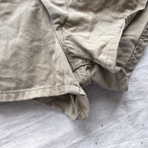 May include: Close-up of khaki-colored shorts made of a textured fabric. The shorts have visible seams and a folded hem. The fabric appears to be slightly wrinkled, suggesting a casual style. The shorts are likely for everyday wear.
