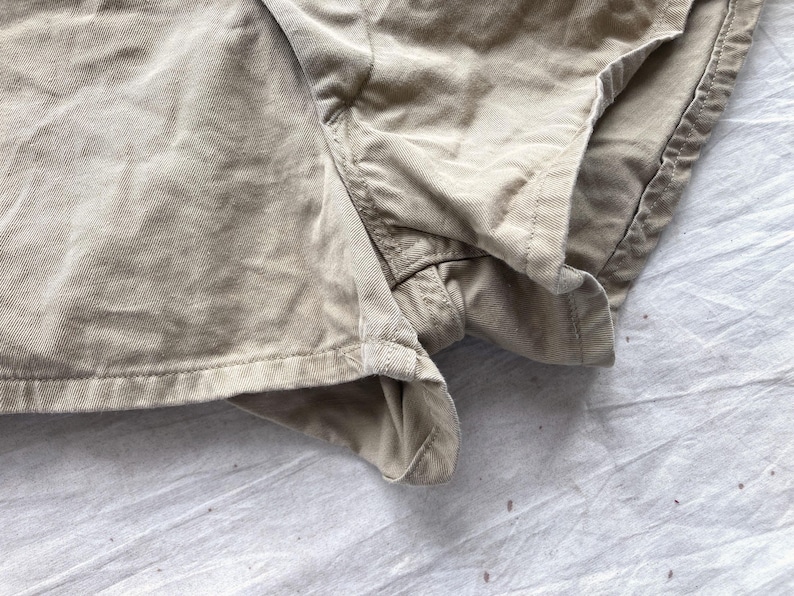 May include: Close-up of khaki-colored shorts made of a textured fabric. The shorts have visible seams and a folded hem. The fabric appears to be slightly wrinkled, suggesting a casual style. The shorts are likely for everyday wear.