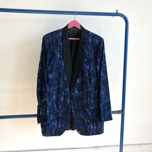 May include: A blazer featuring a dark blue and black abstract pattern with a solid black shawl collar. The garment has long sleeves, a single front button closure, and a structured silhouette, hanging on a pink hanger against a plain wall.