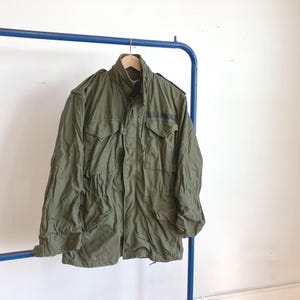 May include: An olive green military jacket with the text "U.S. AIR FORCE" on the upper right chest. The jacket has multiple pockets, a high collar, and is hanging on a wooden hanger from a blue metal rack. The jacket is made of a durable fabric.