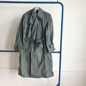 May include: A grey trench coat with a double-breasted design, belt, and button closures. The coat is hanging on a blue metal clothing rack against a white wall. The coat has a classic, utilitarian style.