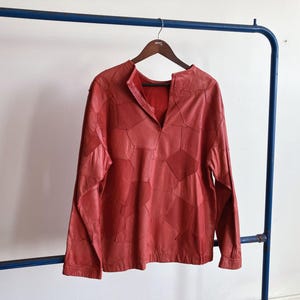 Puede incluir: Una camisa de cuero roja de manga larga con un diseño de patchwork geométrico. La camisa tiene cuello en V y está colgada de una percha de madera. La prenda se exhibe sobre un fondo blanco.