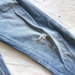 Peut inclure: Gros plan sur un jean en denim bleu clair avec un aspect us&eacute; et vieilli. Le jean pr&eacute;sente une d&eacute;chirure visible au niveau du genou, mettant en valeur les bords effiloch&eacute;s du tissu. Les coutures sont visibles le long des coutures.