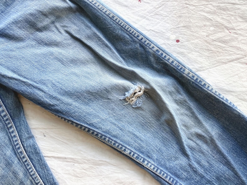 Peut inclure: Gros plan sur un jean en denim bleu clair avec un aspect us&eacute; et vieilli. Le jean pr&eacute;sente une d&eacute;chirure visible au niveau du genou, mettant en valeur les bords effiloch&eacute;s du tissu. Les coutures sont visibles le long des coutures.