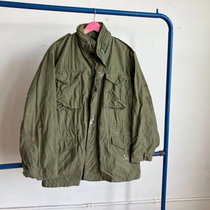 May include: An olive green military-style jacket with multiple front pockets and a high collar. The jacket is hanging on a blue metal rack with a pink hanger. There are white paint splatters on the jacket.