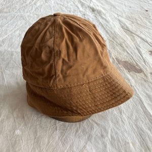 May include: A brown corduroy bucket hat with a rounded crown and a short, structured brim. The hat is a solid color and appears to be made of a soft, textured fabric. The hat is sitting on a light-colored surface.