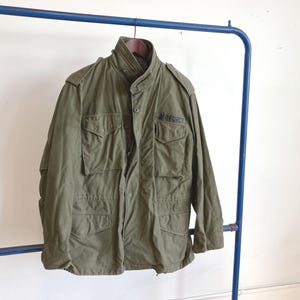 May include: Olive green military-style jacket with multiple pockets and the text "AIR FORCE" on the chest. The jacket is hanging on a blue metal rack against a white wall. The jacket has a button-up front and a high collar.