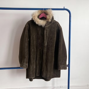 Op de afbeelding: Een donkerbruine jas met een met imitatiebont gevoerde capuchon hangt aan een roze hanger. De jas is gemaakt van een zacht, pluche materiaal en heeft een lange lengte. De jas hangt aan een blauw metalen kledingrek.