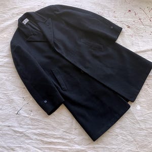 Könnte beinhalten: Ein langer, dunkelmarineblauer Mantel mit klassischem Design. Der Mantel hat ein fallendes Revers, zwei Vordertaschen und einen Einzelknopfverschluss. Der Mantel ist aus einem weichen Material, möglicherweise Wolle.