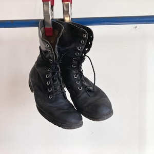 May include: A pair of black leather lace-up boots with silver-toned metal eyelets and black laces. The boots feature a high-top design with a rounded toe and a sturdy sole, currently suspended from a blue horizontal bar by two red-handled clamps.