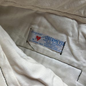 K&ouml;nnte beinhalten: Nahaufnahme eines Etiketts in einem Kleidungsst&uuml;ck. Das Etikett tr&auml;gt die Aufschrift "Original UBERMEYER MADE IN BAVARIA GERMANY 100% COTTON". Ein rotes Herz ist auf dem Etikett zu sehen. Das Kleidungsst&uuml;ck ist wei&szlig;.