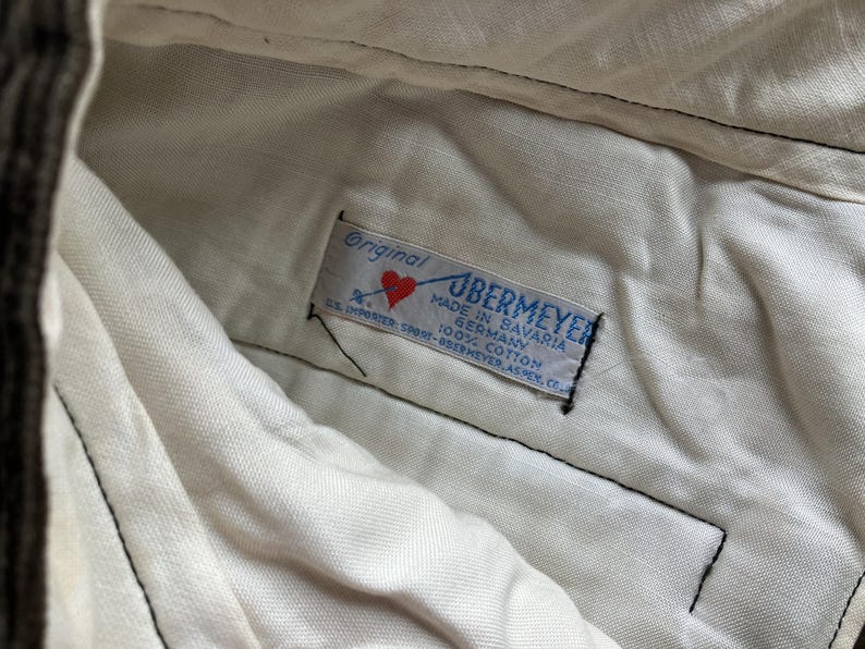 K&ouml;nnte beinhalten: Nahaufnahme eines Etiketts in einem Kleidungsst&uuml;ck. Das Etikett tr&auml;gt die Aufschrift "Original UBERMEYER MADE IN BAVARIA GERMANY 100% COTTON". Ein rotes Herz ist auf dem Etikett zu sehen. Das Kleidungsst&uuml;ck ist wei&szlig;.