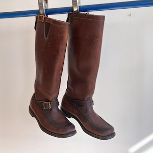 Puede incluir: Un par de botas altas de cuero marrón con puntera estilo mocasín, una correa con hebilla de tono latón en el tobillo y una segunda correa con hebilla en la parte superior de la caña. Las botas tienen un acabado en color marrón oscuro.