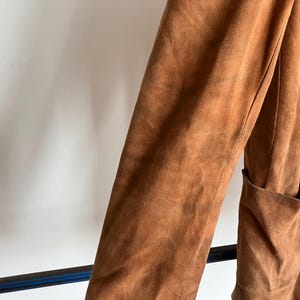 Może przedstawiać: Zbliżenie brązowej kurtki zamszowej. Kurtka posiada długi rękaw i kieszeń. Materiał zamszowy ma teksturowany wygląd. Kurtka ma ciepły, ziemisty odcień.
