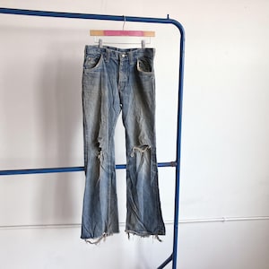 Puede incluir: Unos pantalones vaqueros de mezclilla azul con un lavado desteñido, cierre de botones y roturas prominentes en ambas rodillas. Las aberturas de las piernas están deshilachadas y desgastadas, mostrando un corte relajado con bolsillos clásicos.