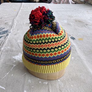 May include: A yellow knit beanie with a colorful geometric pattern and a red, blue, and black pom-pom on top.