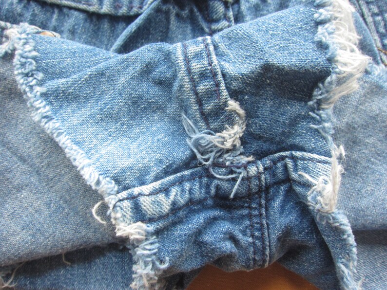 Op de afbeelding: Close-up van een paar blauwe denim shorts met rafelige randen. De stof is verweerd en heeft een verbleekte look.