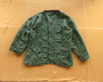Puede incluir: Chaqueta acolchada verde oliva con un patrón de diamantes. La chaqueta tiene cuello en V y mangas largas. La chaqueta está colocada sobre una superficie de color beige.