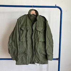 Op de afbeelding: Een olijfgroene jas in militaire stijl met meerdere voorzakken en een knoopsluiting. De jas hangt aan een houten hanger aan een blauw metalen kledingrek. De jas heeft een hoge kraag.