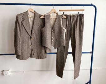 May include: A three-piece suit in a brown tweed fabric hangs on a blue rack. The suit includes a blazer with gold buttons, a vest, and matching trousers. The garments are displayed on wooden hangers against a white wall.