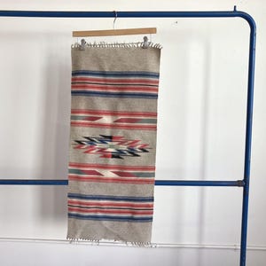 May include: A rectangular woven wall hanging with a beige background and horizontal stripes in red, blue, and gray. The center features a geometric design in red, black, and blue. The edges have a fringe.