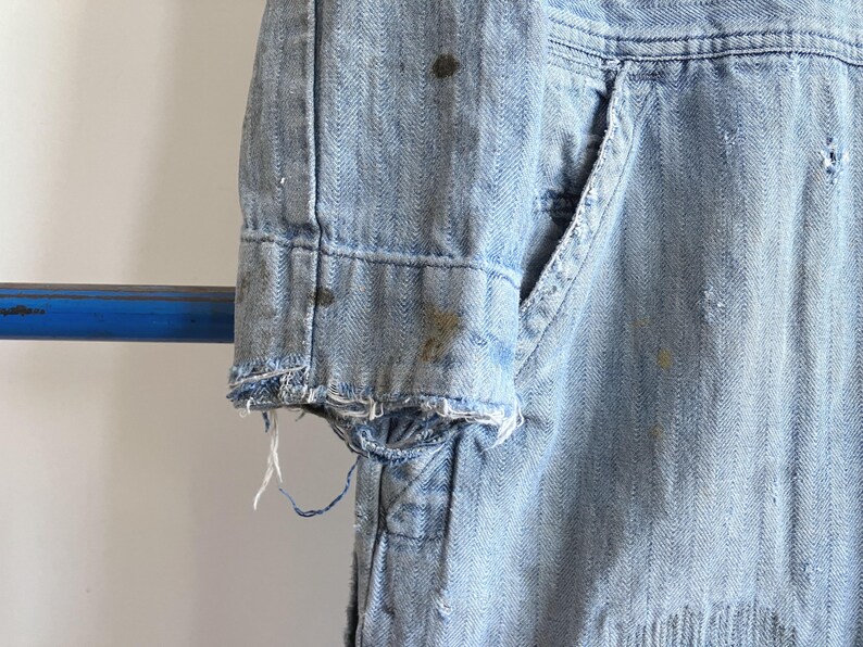 May include: Close-up of a light blue denim jacket sleeve. The sleeve has a frayed edge and visible wear and tear, including paint splatters. The jacket features a pocket and visible stitching details.