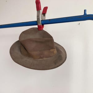 May include: A weathered, taupe-coloured fedora hat with a creased crown and a wide brim. The hat is secured to a blue metal bar by a red and silver spring clamp. The fabric shows signs of wear and discolouration, particularly around the hat band area.