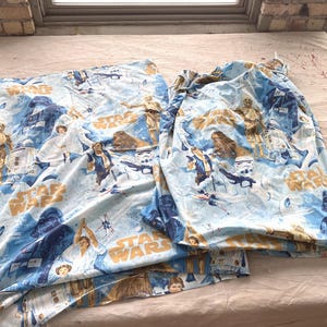 Może przedstawiać: Jasnoniebieska tkanina o tematyce Gwiezdnych Wojen z różnymi postaciami, takimi jak Darth Vader, Luke Skywalker i Chewbacca. Napis "STAR WARS" jest wydrukowany w kolorze złotym. Tkanina jest prawdopodobnie przeznaczona na pościel lub zasłony.