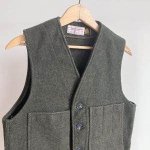 May include: A dark olive green wool vest featuring a V-neckline, a button-front closure with three dark buttons, and two large front patch pockets. A white rectangular label with black text is sewn inside the neckline of the garment.