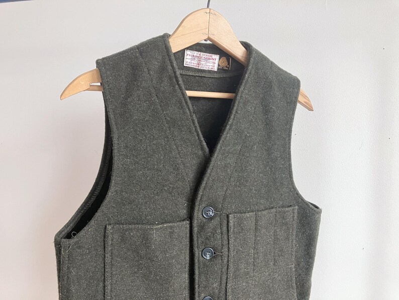 May include: A dark olive green wool vest featuring a V-neckline, a button-front closure with three dark buttons, and two large front patch pockets. A white rectangular label with black text is sewn inside the neckline of the garment.