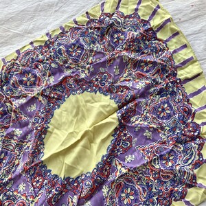 May include: A square scarf with a paisley design. The scarf features a central oval of pale yellow, surrounded by a border of intricate purple, red, and blue paisley patterns. The edges are pale yellow with purple stripes.