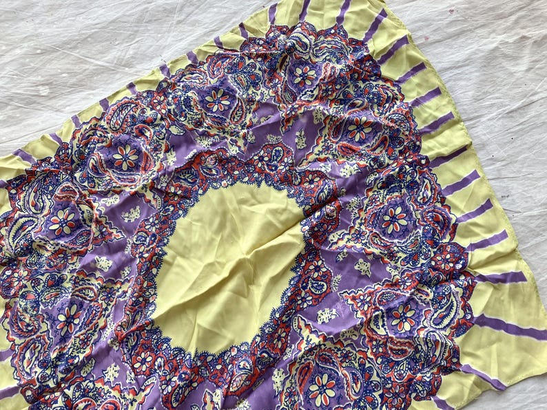 May include: A square scarf with a paisley design. The scarf features a central oval of pale yellow, surrounded by a border of intricate purple, red, and blue paisley patterns. The edges are pale yellow with purple stripes.