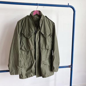 May include: An olive green military-style jacket with multiple front pockets and a button-up closure. The jacket is hanging on a pink hanger attached to a blue metal clothing rack. The jacket has a high collar and long sleeves.