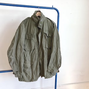 Op de afbeelding: Olijfgroene jas in militaire stijl met meerdere zakken en een hoge kraag, hangend aan een blauw metalen rek. De jas heeft een knoopsluiting en een taille met trekkoord. De stof lijkt een duurzaam, licht versleten materiaal te zijn.