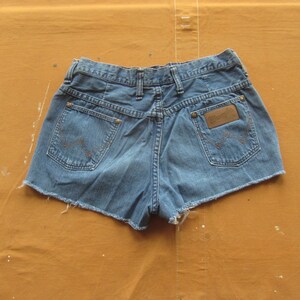 Op de afbeelding: Een blauwe denim short met een rafelige zoom. De short heeft een bruin leren patch met het woord "Wrangler" op de rechter achterzak.