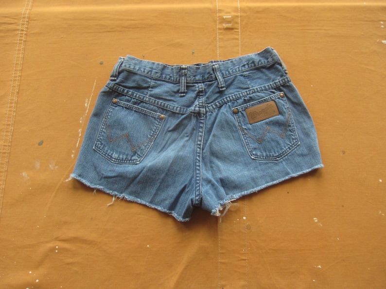 Op de afbeelding: Een blauwe denim short met een rafelige zoom. De short heeft een bruin leren patch met het woord "Wrangler" op de rechter achterzak.