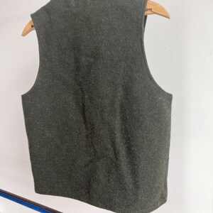 May include: A sleeveless vest in a dark olive green color with a textured, felted appearance. The garment is displayed on a wooden hanger, showing a simple, straight-cut design with wide arm openings and a rounded neckline, viewed from the back.
