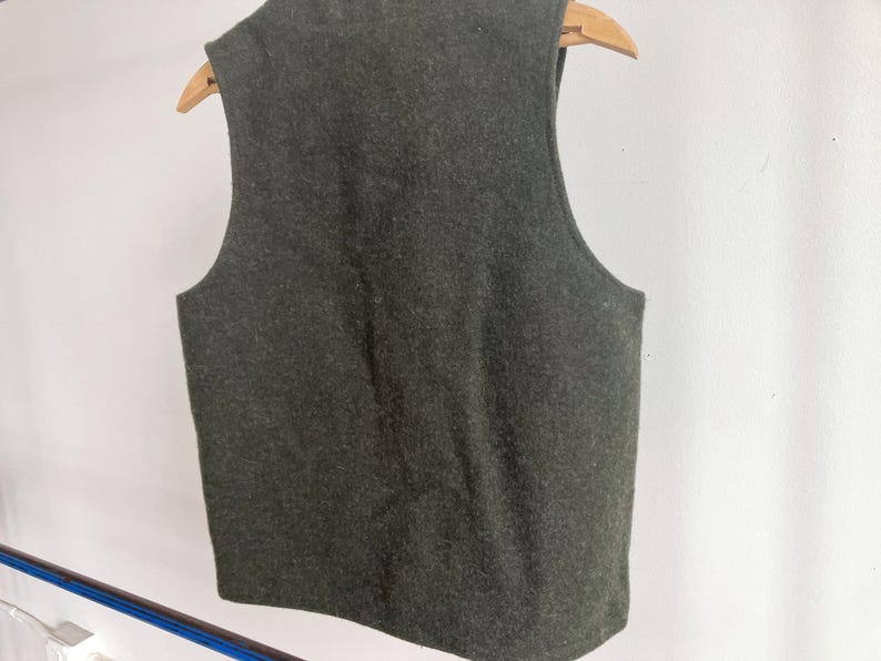 May include: A sleeveless vest in a dark olive green color with a textured, felted appearance. The garment is displayed on a wooden hanger, showing a simple, straight-cut design with wide arm openings and a rounded neckline, viewed from the back.