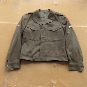 Puede incluir: Una chaqueta militar verde con cierre de botones y dos bolsillos de solapa. La chaqueta tiene un parche en la manga izquierda con un diseño rojo y blanco.