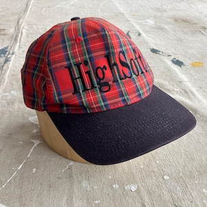May include: A red, blue, and green tartan baseball cap with a navy blue peak. The text "High Soft" is embroidered in black across the front. The cap is displayed on a tan form.