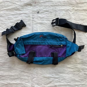 May include: A teal and purple fanny pack with black straps and buckles. The pack has a zippered pocket and the brand name "Lowe" in red. The fanny pack is designed to be worn around the waist and is made of a durable material.