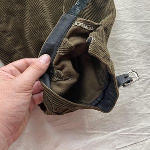 K&ouml;nnte beinhalten: Nahaufnahme eines olivgr&uuml;nen Cordhosenbeins mit einem schwarzen Lederriemen und silberner Schnalle. Die Hose hat ein strukturiertes, geripptes Design und ein dunkles Innenfutter.