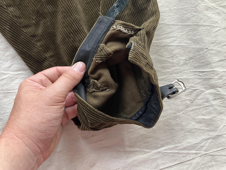 K&ouml;nnte beinhalten: Nahaufnahme eines olivgr&uuml;nen Cordhosenbeins mit einem schwarzen Lederriemen und silberner Schnalle. Die Hose hat ein strukturiertes, geripptes Design und ein dunkles Innenfutter.