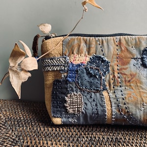 May include: A rectangular zippered pouch featuring a patchwork design of tan burlap and various blue denim fabric scraps. The surface is decorated with intricate hand-stitched embroidery patterns, including running stitches and cross-stitch details.